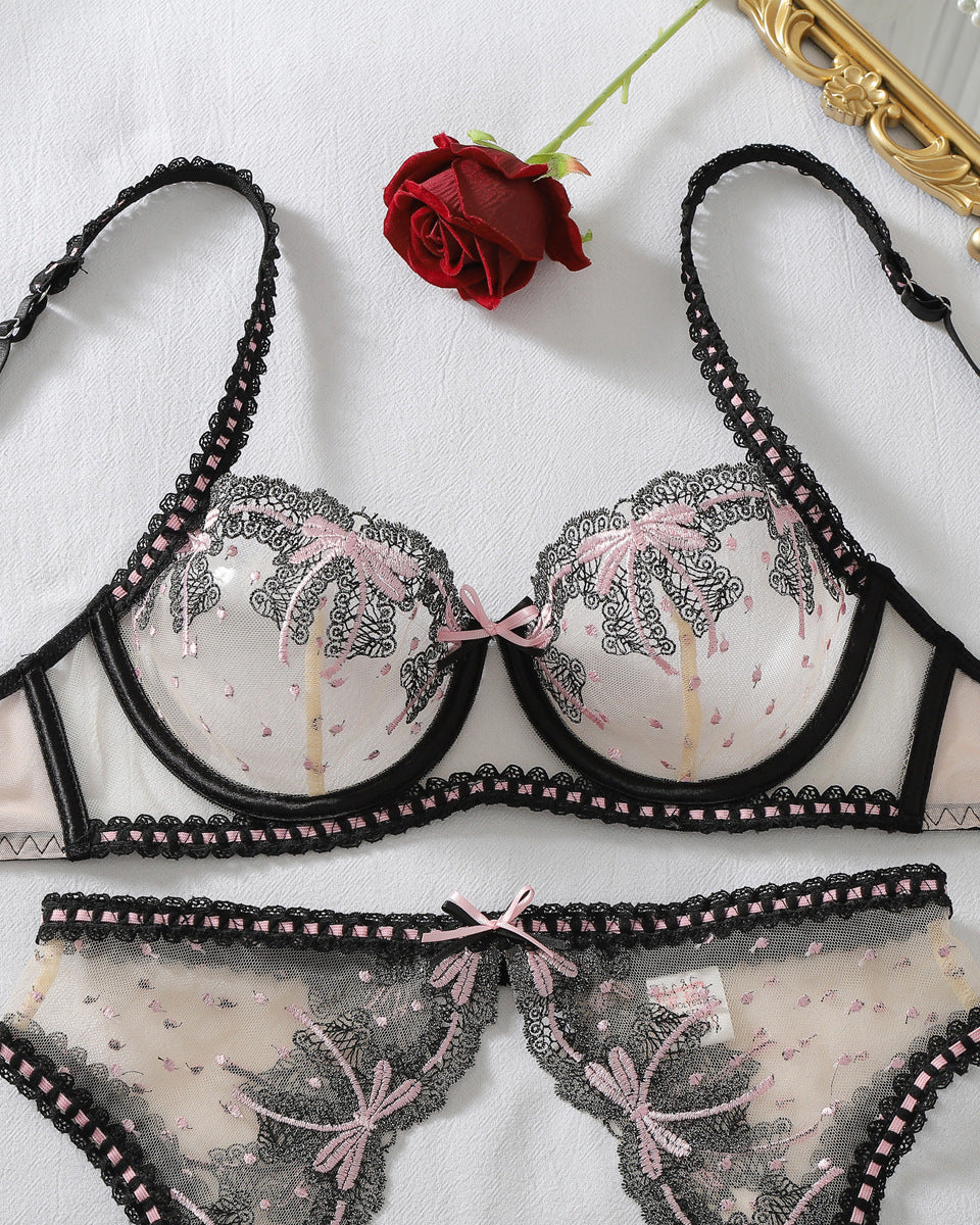A two-piece lingerie set featuring a sheer bra and matching panties with black lace, pink bow accents, and a tropical palm tree print, styled on a white fabric background with a red rose and a gold mirror edge.
